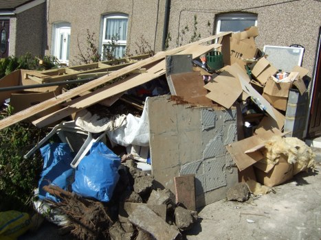 A large pile of mixed household waste and debris is stacked on a pavement outside a residential building, likely for rubbish removal. The mound includes broken wooden planks, cardboard boxes, and parts of a discarded appliance or furniture, with some corrugated cardboard and paper visible. There are also fabric materials, a black plastic bag, and rubble composed of small concrete fragments and dirt. The waste is haphazardly piled, partially leaning against the wall of a grey stone building with white-framed windows. The scene appears to be in a residential area, possibly in or near Seven Sisters, London. The overall environment shows clear daylight and urban surroundings, with the waste occupying a section of the sidewalk or driveway. The image reflects a typical arrangement of rubbish awaiting collection, representative of services provided by Business Waste Removal Seven Sisters for effective waste clearance and disposal.