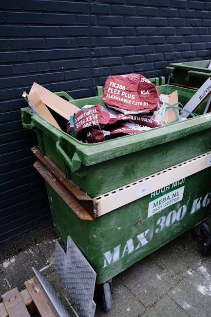 A large green waste bin filled with red and black bags of garden or DIY waste, placed on a paved surface against a dark blue brick wall. The bags appear to contain soil, mulch, or similar bulky garden